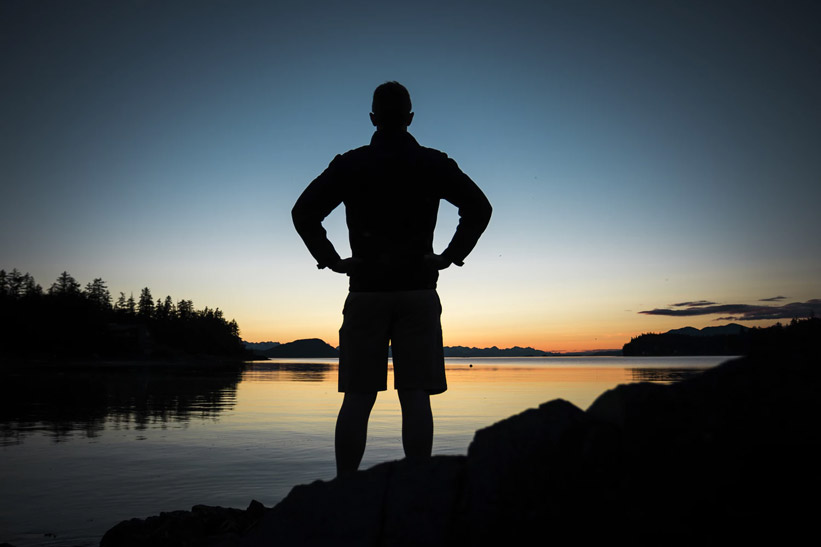 A man standing on the shore of a lake at sunset.